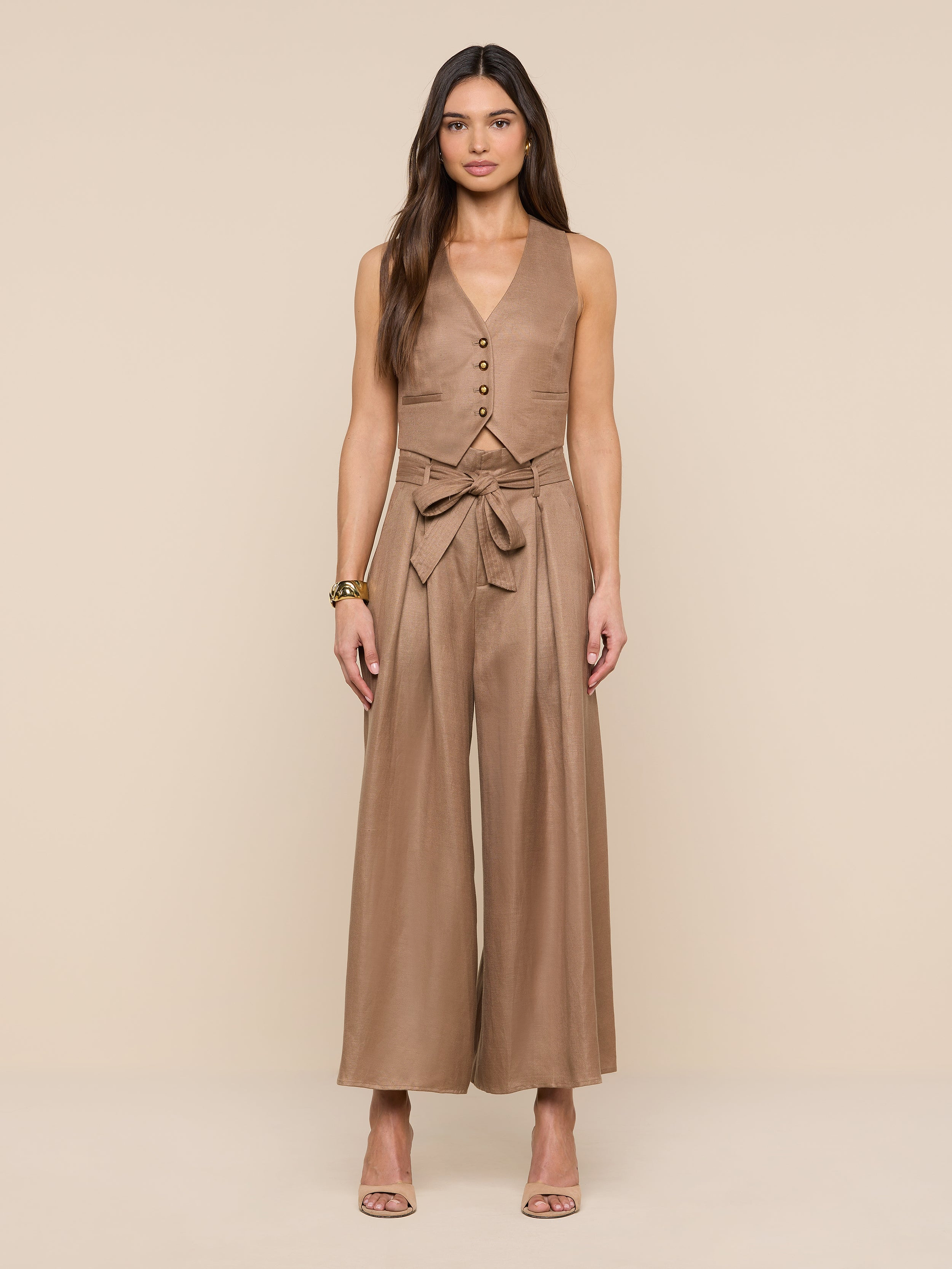 A woman stands facing forward, wearing the Lowen Linen-Blend Pant in tan, styled with a matching sleeveless top and a tied belt. She has long hair, neutral heels, and a bracelet, posing against a plain white background.