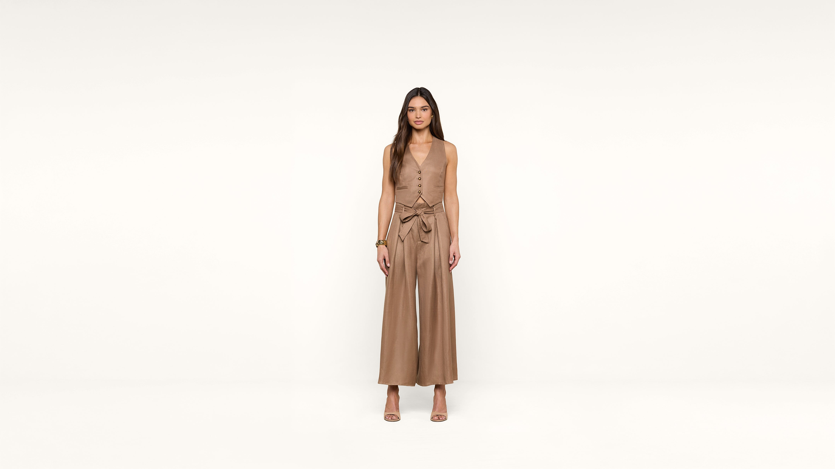A woman stands facing forward against a plain white background, wearing a sleeveless, tan jumpsuit with wide legs and a tied waist, along with heeled sandals.