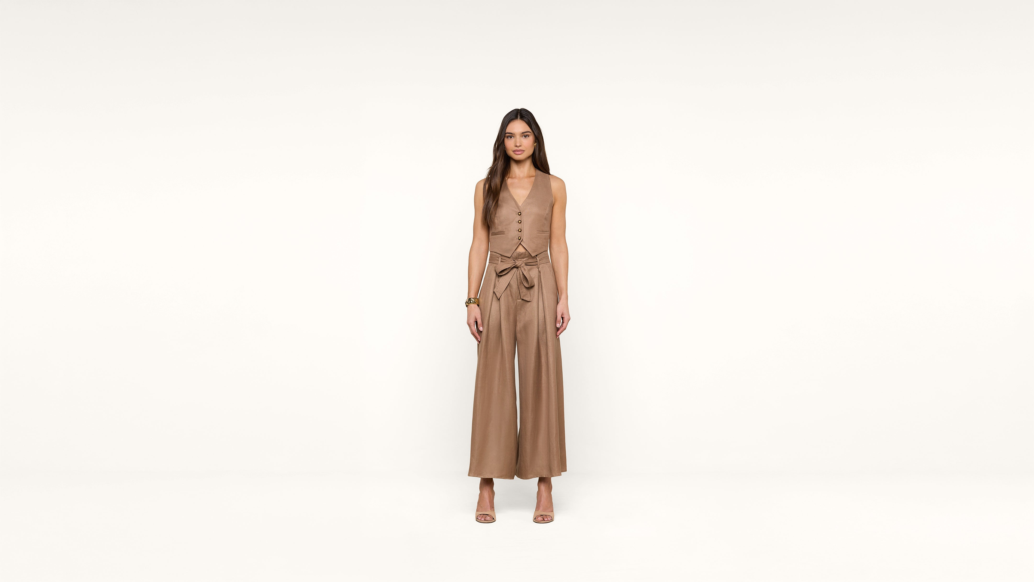 A woman stands facing forward against a plain white background, wearing a sleeveless, tan jumpsuit with wide legs and a tied waist, along with heeled sandals.