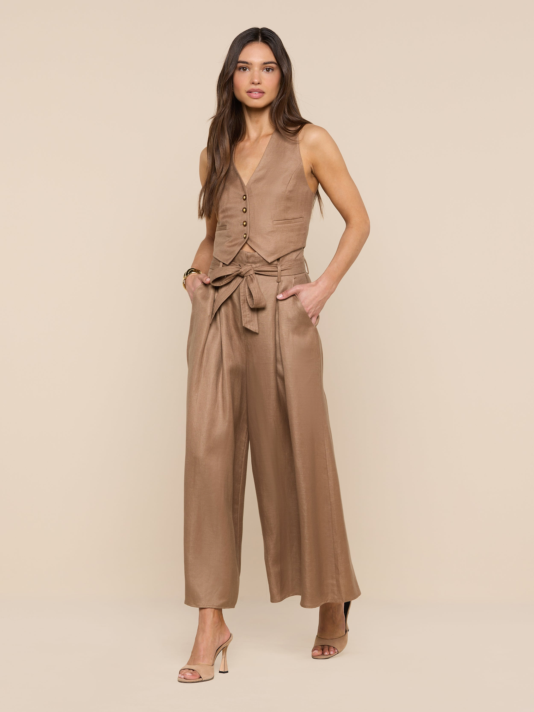 A woman stands against a white background, hands in her pockets, wearing tan open-toe heels and the Lowen Linen-Blend Pant—a wide-leg, cropped style with a tied belt—paired with a coordinating tan sleeveless buttoned vest.