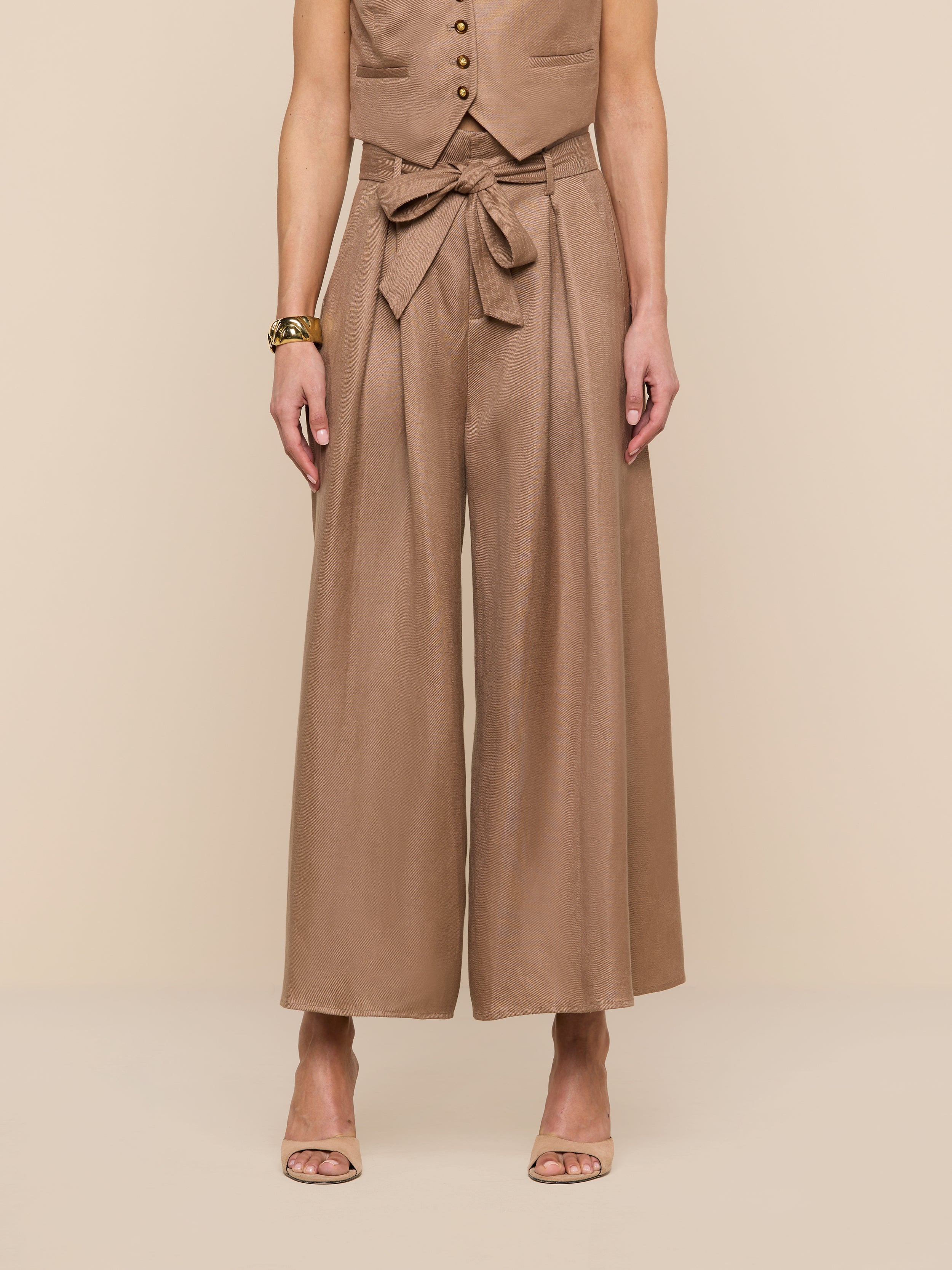 A person wearing a sleeveless tan vest, the Lowen Linen-Blend Pant featuring a wide cropped leg and tied waist, and tan heeled sandals stands against a plain white background.
