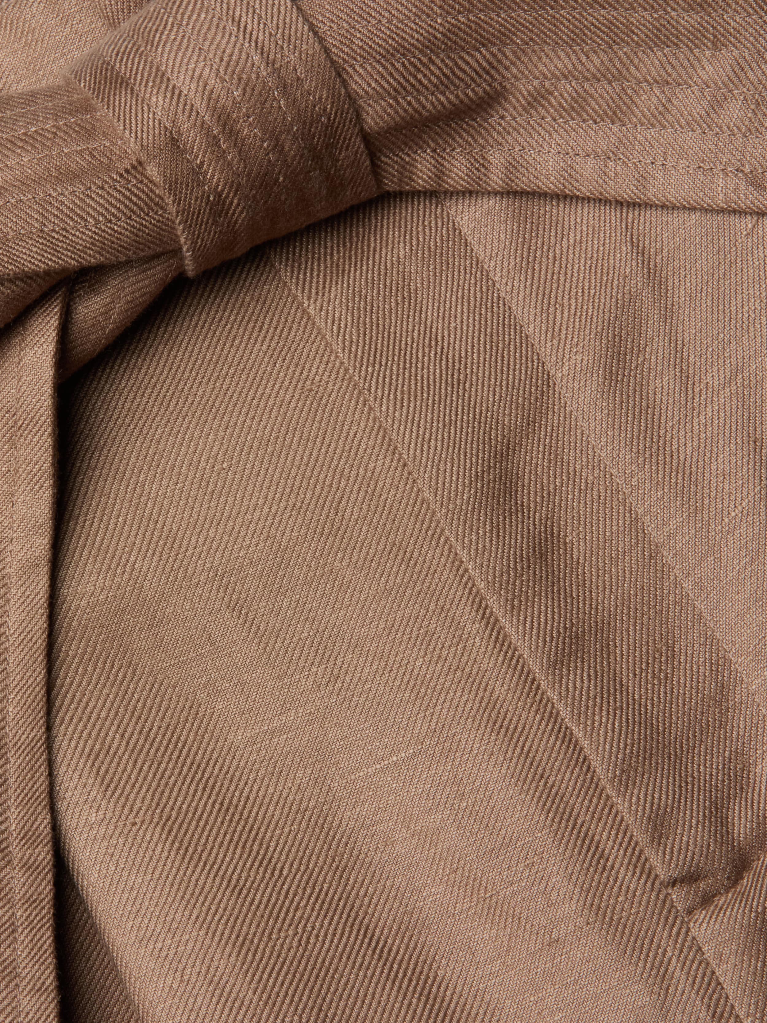 Close-up of brown linen-blend fabric with a visible twill weave pattern, highlighting a neatly sewn seam and a fabric fold or belt crossing over itself—details that reflect the refined tailoring of the Lowen Linen-Blend Pant with its wide cropped leg.