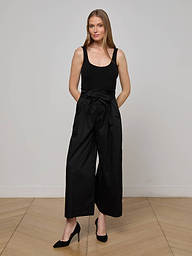 A woman stands against a plain white background wearing a sleeveless black top, black heels, and the Lowen Pant—wide leg black poplin pants featuring a waist tie.