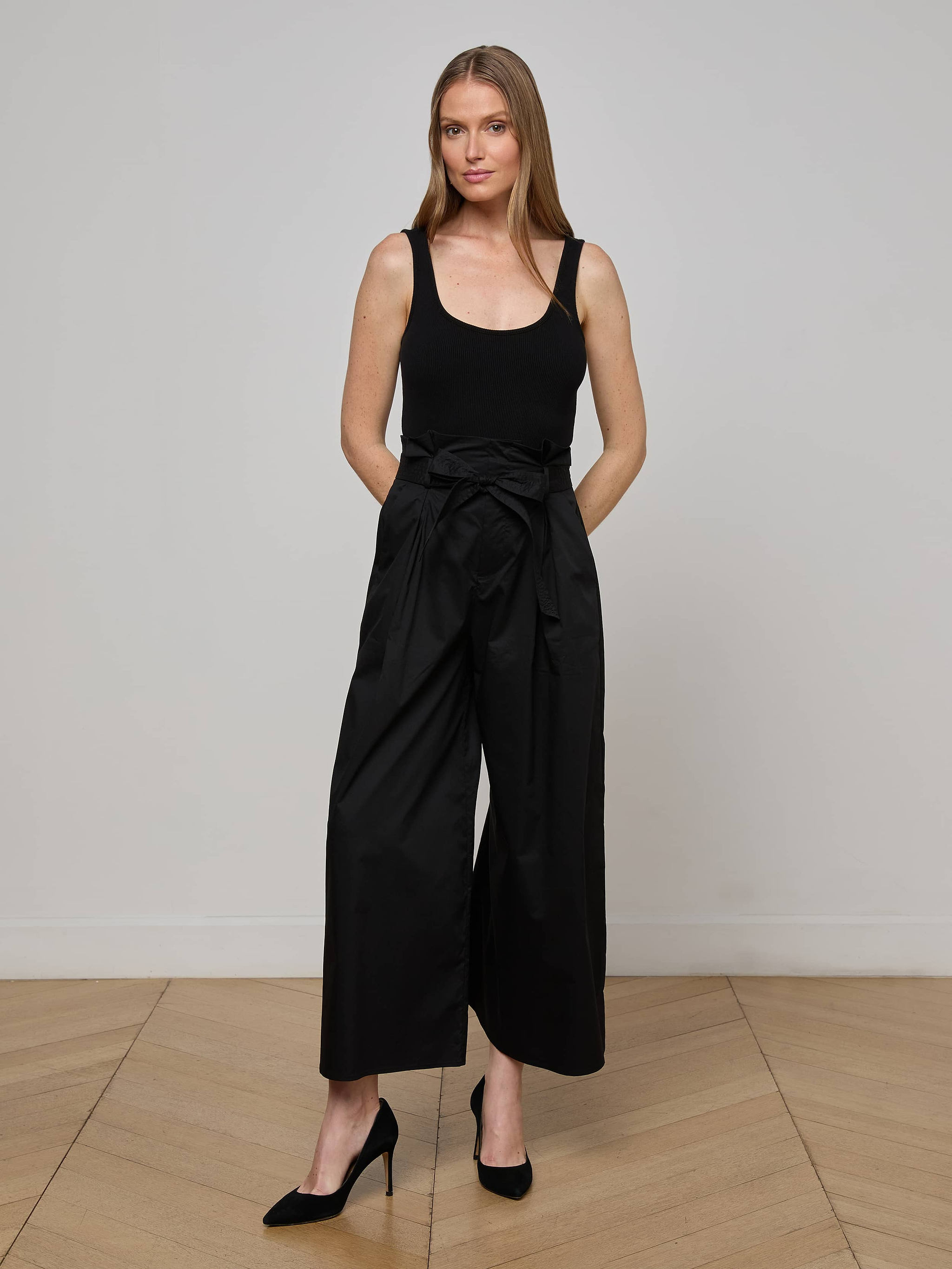 A woman stands against a plain white background wearing a sleeveless black top, black heels, and the Lowen Pant—wide leg black poplin pants featuring a waist tie.