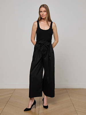 A woman stands indoors against a light wall, wearing the L'AGENCE Lowen Pant—high rise wide leg cropped black poplin pants with a tied belt—paired with a black sleeveless top and heels. She has long straight hair and smiles slightly.