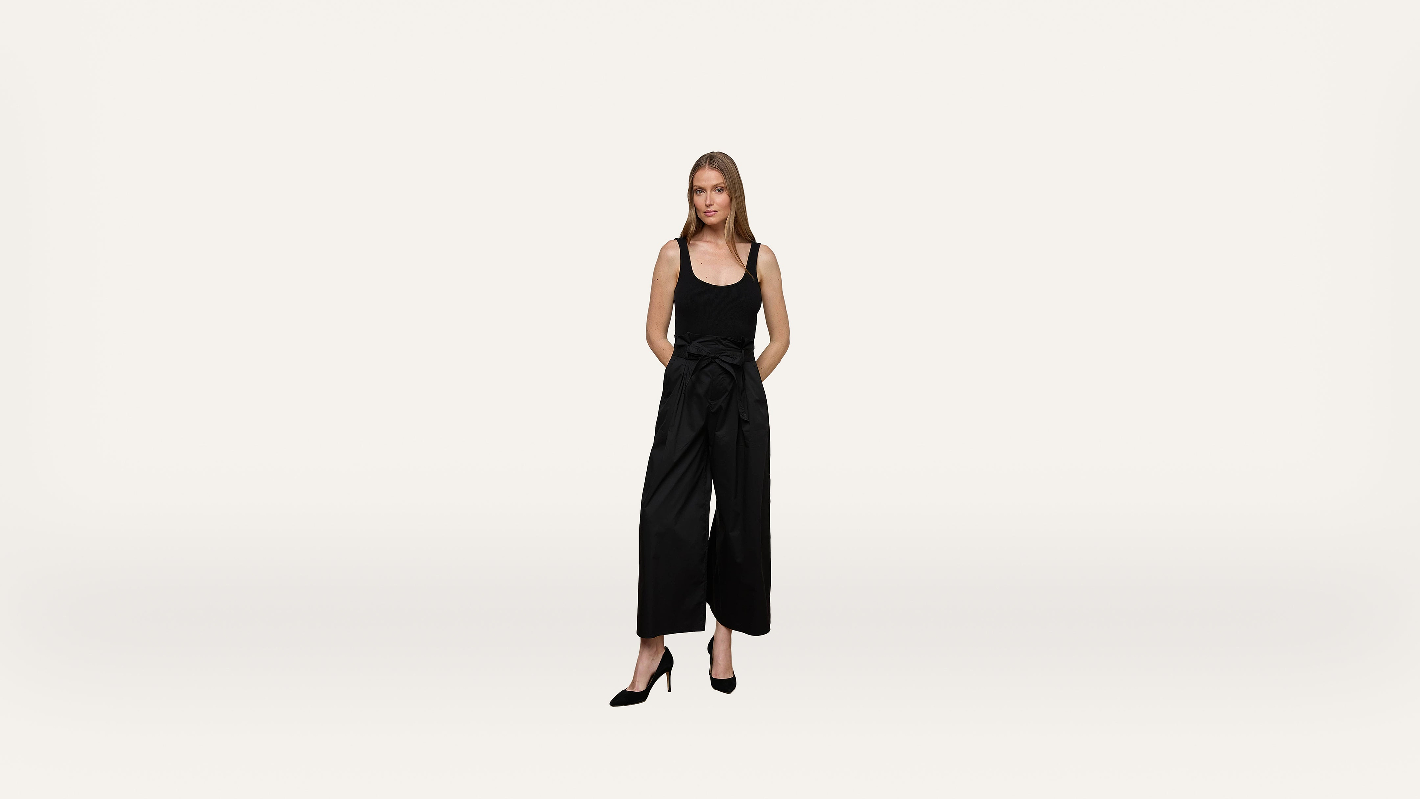A woman in a black sleeveless top, wide-leg black pants, and black heels stands with hands behind her back against a plain white background.
