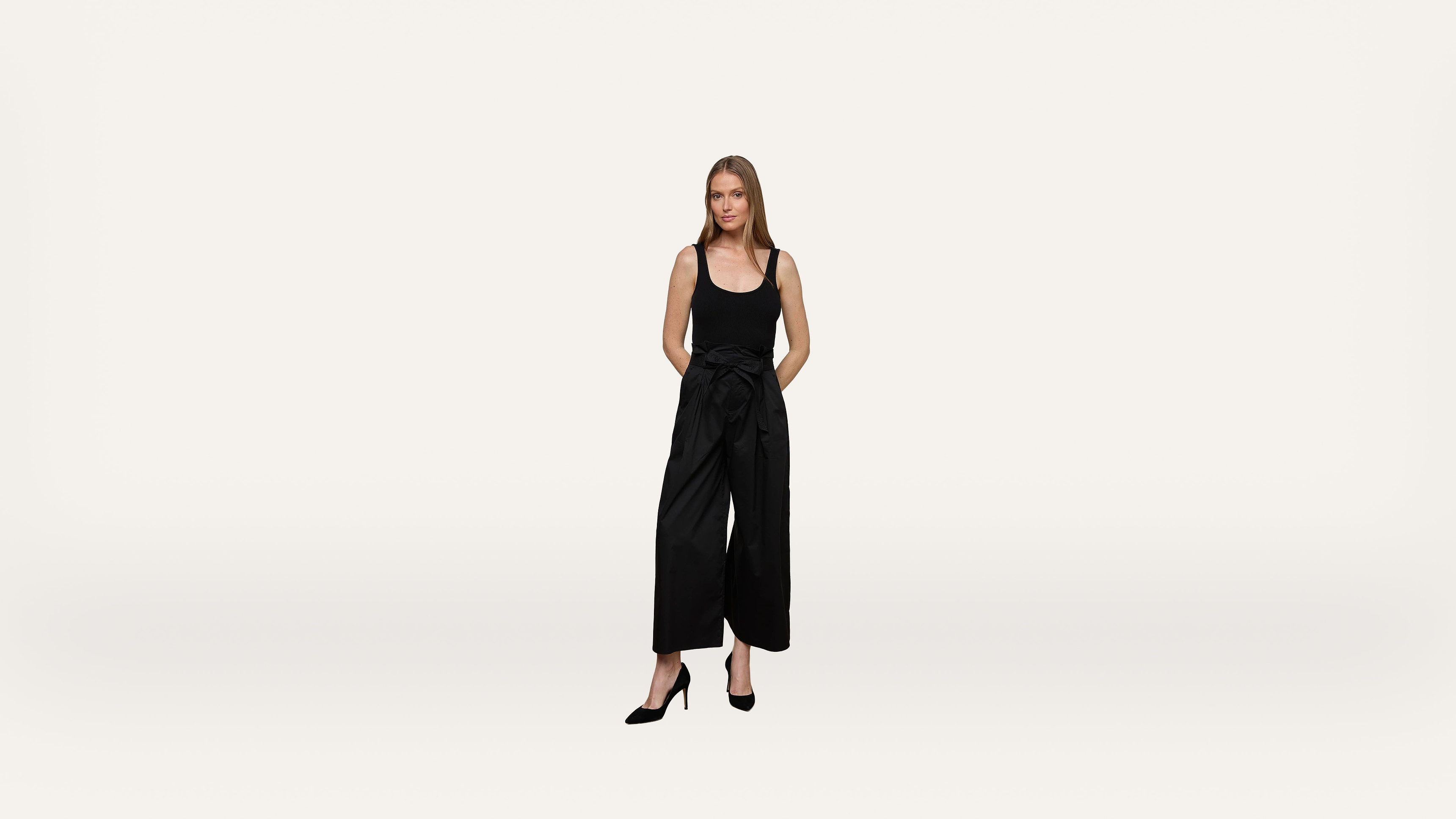 A woman in a black sleeveless top, wide-leg black pants, and black heels stands with hands behind her back against a plain white background.