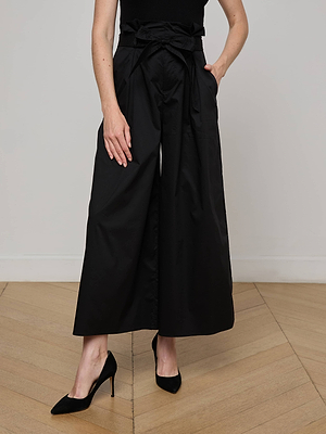 A woman stands on a wooden floor wearing the L'AGENCE Lowen Pant—wide-leg black poplin pants with a bow belt—paired with a black sleeveless top and pointed-toe heels. Her left hand is in her pocket.