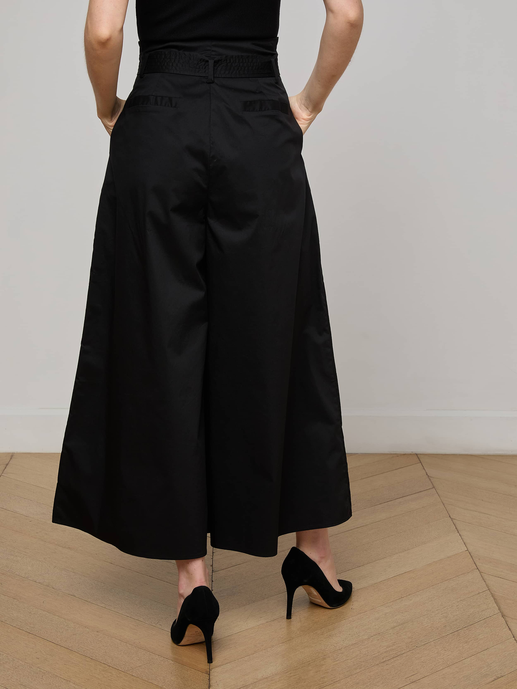 Wearing the Lowen Pant—high rise, wide-leg black poplin pants—with black high heels, the person is shown from the back against a plain light background. Their upper body is partially visible and their arms rest relaxed at their sides.