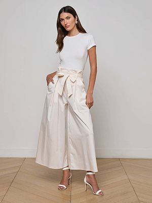 A woman stands indoors wearing the L'AGENCE Lowen Pant, a high-rise paperbag waist style with a large bow, paired with a fitted white top and white heeled sandals. The setting features light walls and wooden flooring.