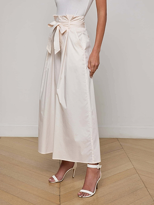 A woman stands on a light wooden floor against a plain wall, wearing a white sleeveless top, L'AGENCE Lowen Pant—vintage white high-rise paperbag waist pants with a tied belt—and white heeled sandals.