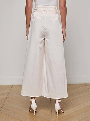 A person stands facing away, wearing the L'AGENCE Lowen Pant, a high-rise paperbag waist style in light pink, paired with white heeled sandals on a wooden floor against a plain light-colored wall.