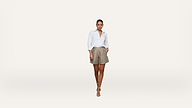 A woman stands against a plain white background, wearing a white long-sleeve shirt, tan shorts, and beige sandals. She has one hand in her pocket and looks confidently at the camera.