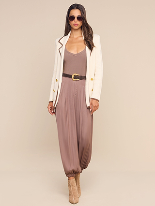 A woman wearing sunglasses, a white blazer, the Palmer Satin Pant in high rise mauve cinched with a belt, and beige heeled boots stands facing forward against a plain beige background.