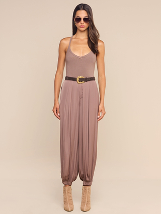 A woman wearing sunglasses, tan heeled boots, and the Palmer Satin Pant—a taupe halter jumpsuit with high-rise balloon trousers cinched by a brown belt—stands against a plain beige background.