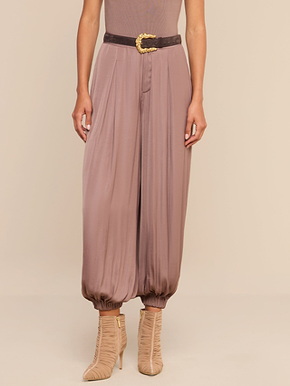 A person wearing the Palmer Satin Pant in a loose, silky mauve style with gathered cuffs, paired with a black belt featuring a gold buckle and beige heeled ruched boots, stands against a neutral background.