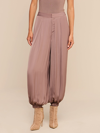 A woman showcases the Palmer Satin Pant—a high-rise, loose-fitting mauve satin trouser designed with pleats and gathered ankles in a chic balloon silhouette. She pairs the look effortlessly with nude, high-heeled ruched boots, standing with her arms relaxed at her sides against a neutral background.