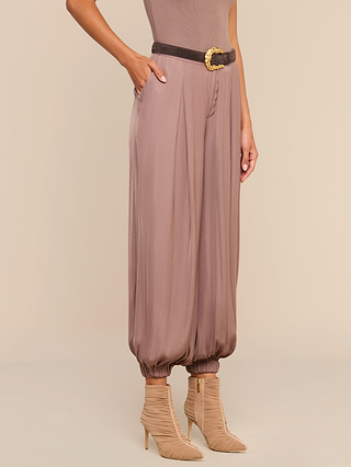 A person wearing the Palmer Satin Pant in taupe, styled high rise with a loose, balloon fit and gathered ankles, paired with a black belt featuring a gold buckle and beige strappy heeled ankle boots, hand in pocket.