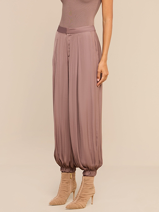 A woman wears loose, high-rise mauve Palmer Satin Pants with gathered ankles and beige heeled boots. Only her lower torso, legs, and part of one arm are visible against a neutral background.