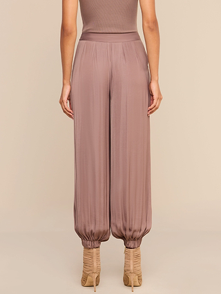 A woman stands facing away, wearing the Palmer Satin Pant in mauve—a high-rise, loose-fitting balloon trouser with gathered cuffs—paired with nude heels against a beige background.