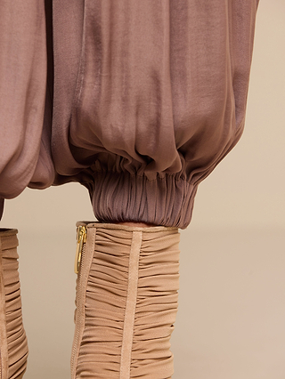 Close-up of the Palmer Satin Pant in taupe, featuring a high-rise fit, ruched detailing, and elastic cuffs for a modern balloon silhouette, paired with beige ruched heeled boots accented by gold zippers.