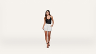 A woman in a black tank top, white shorts, and black heels stands against a plain white background, with one hand in her pocket and looking slightly to the side.