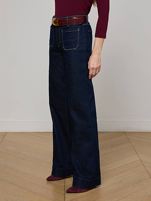 Wearing the Nolan Patch Pocket Jean in a high-rise, dark blue wash with front pockets, burgundy long-sleeve top, brown belt, and burgundy pointed shoes, a person stands on a herringbone wood floor by a light wall.