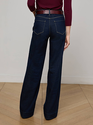 A person stands on a wooden floor facing away from the camera, wearing the Nolan Patch Pocket Jean in dark blue wash paired with a red belt and a long-sleeve burgundy top.