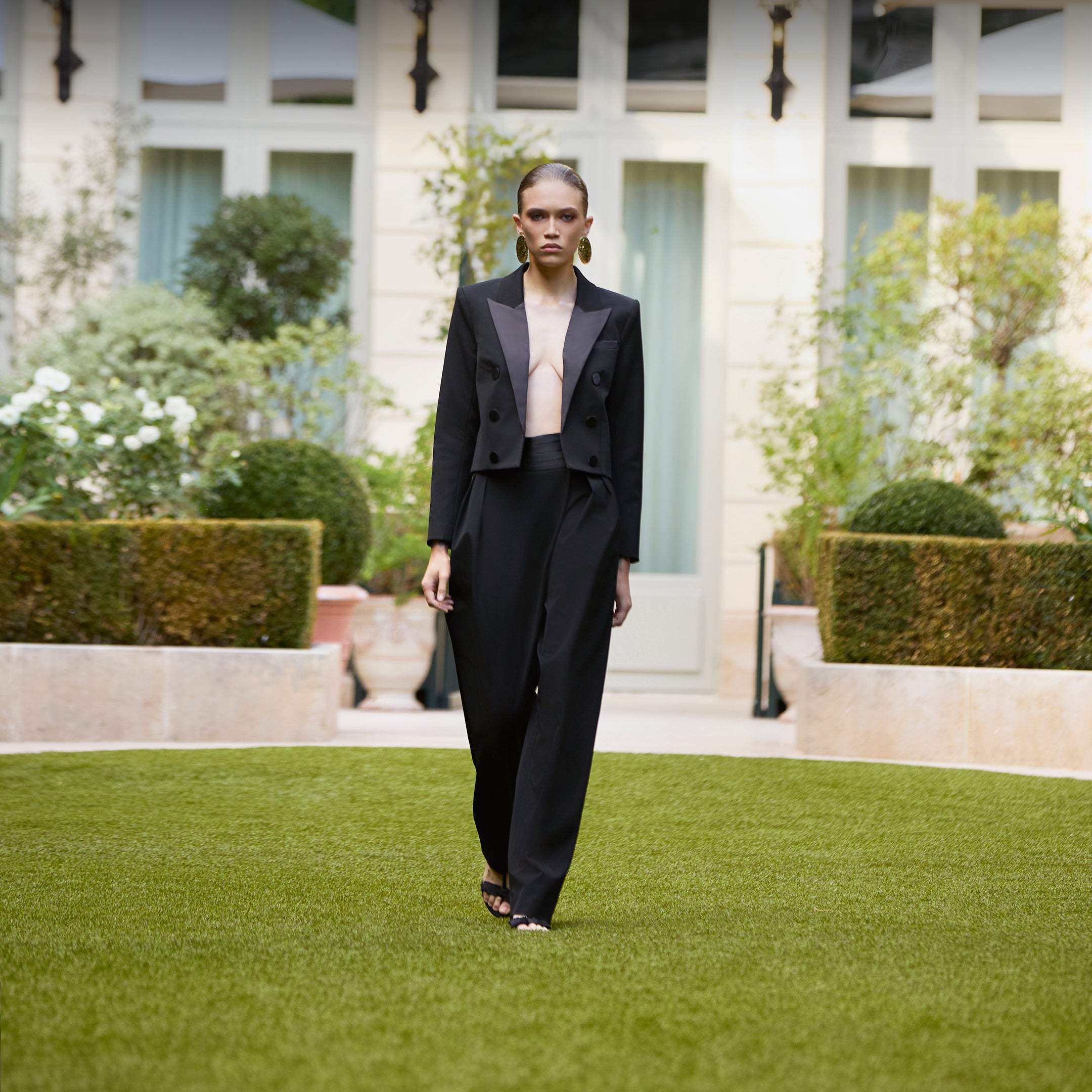 A model walks on a manicured green lawn in front of a grand building, wearing a black cropped blazer with wide lapels and matching wide-leg trousers.