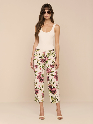A woman in sunglasses, a sleeveless cream bodysuit, Blaine Cropped Stovepipe Jeans with a rose print, and cream heeled sandals stands against a plain beige background.