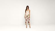 A woman wearing sunglasses, a sleeveless beige top, and white pants with a floral print stands against a plain white background. She is wearing high-heeled sandals.