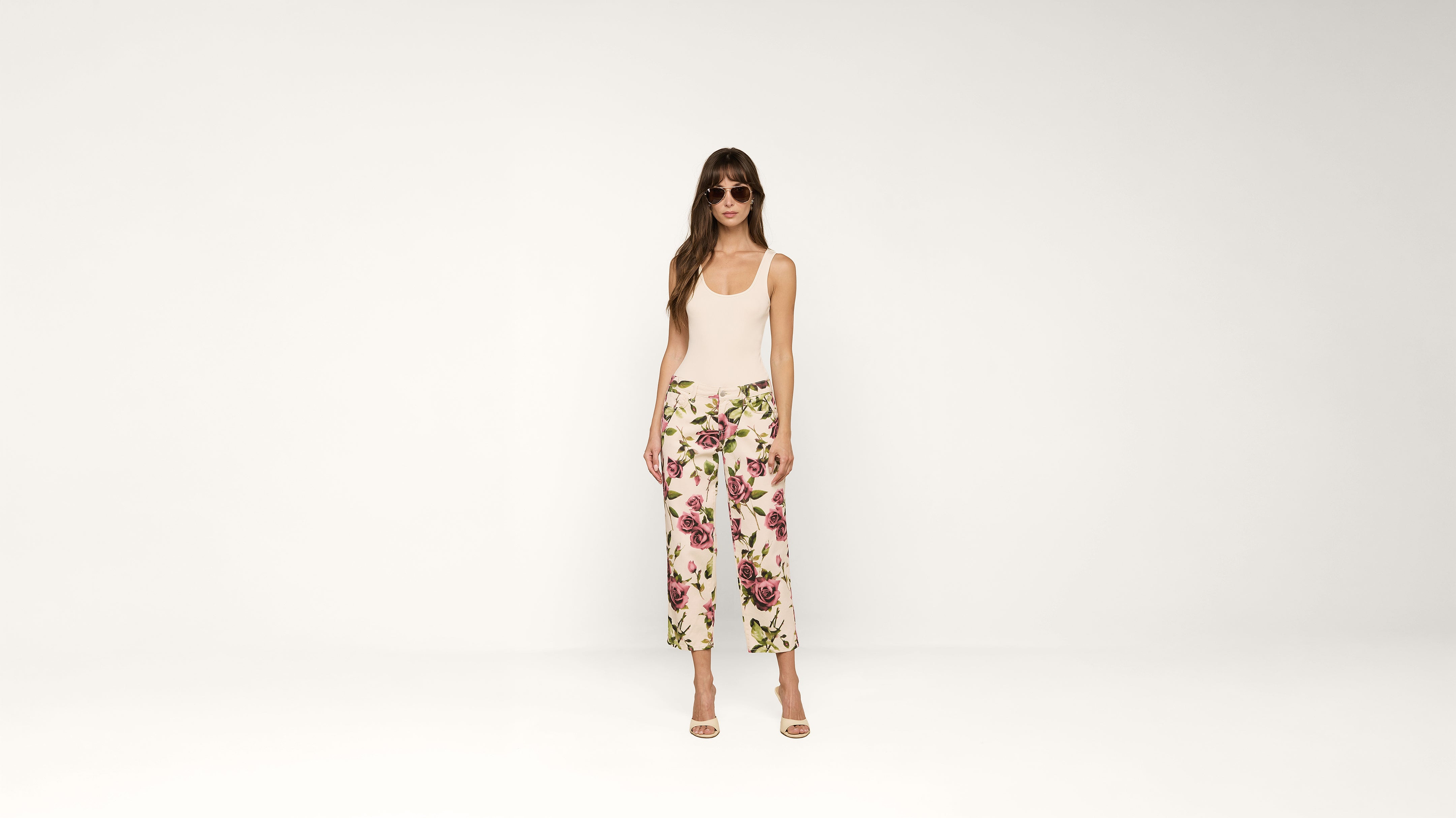 A woman wearing sunglasses, a sleeveless beige top, and white pants with a floral print stands against a plain white background. She is wearing high-heeled sandals.