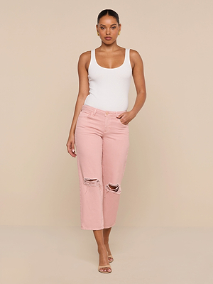 A woman stands against a beige background wearing a white sleeveless top, Blaine Cropped Stovepipe Jean in light pink with ripped details, and open-toe heels, posing with one hand in her pocket and a confident expression.