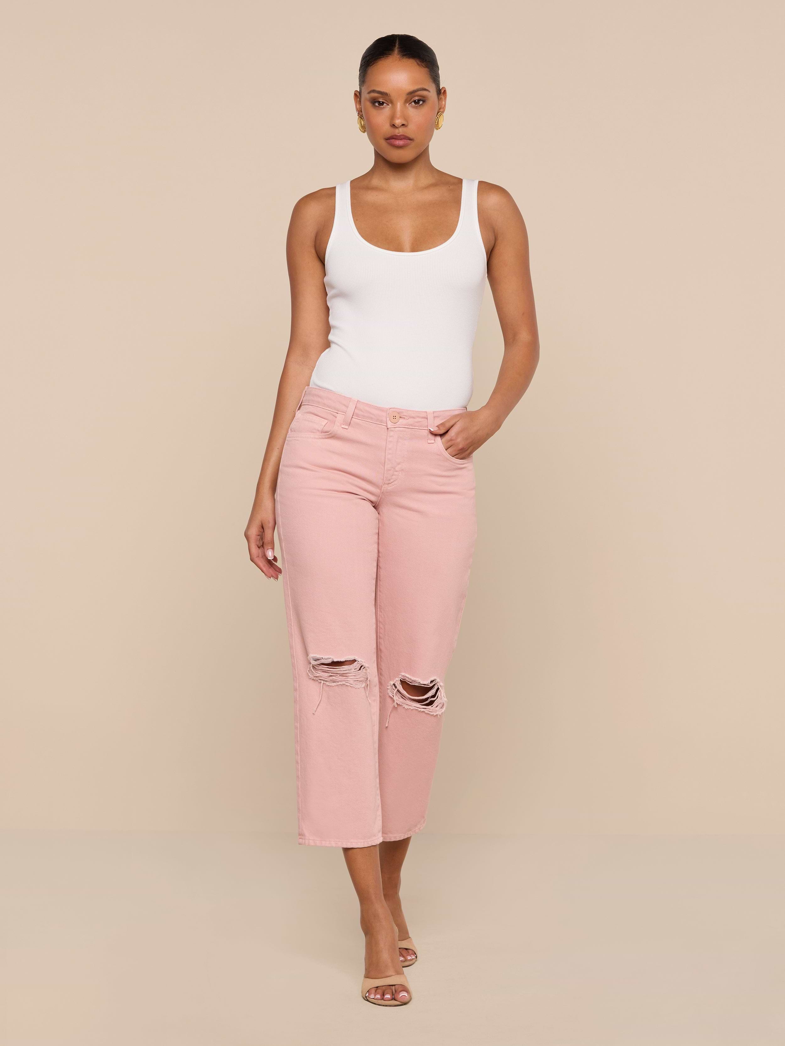 A woman stands against a plain white background wearing a white tank top, the Blaine Cropped Stovepipe Jean in light pink with a straight-leg fit and low rise, and heeled sandals, with one hand in her pocket.