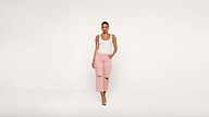 A woman stands against a plain white background, wearing a white tank top, light pink ripped jeans, and sandals, with one hand in her pocket.