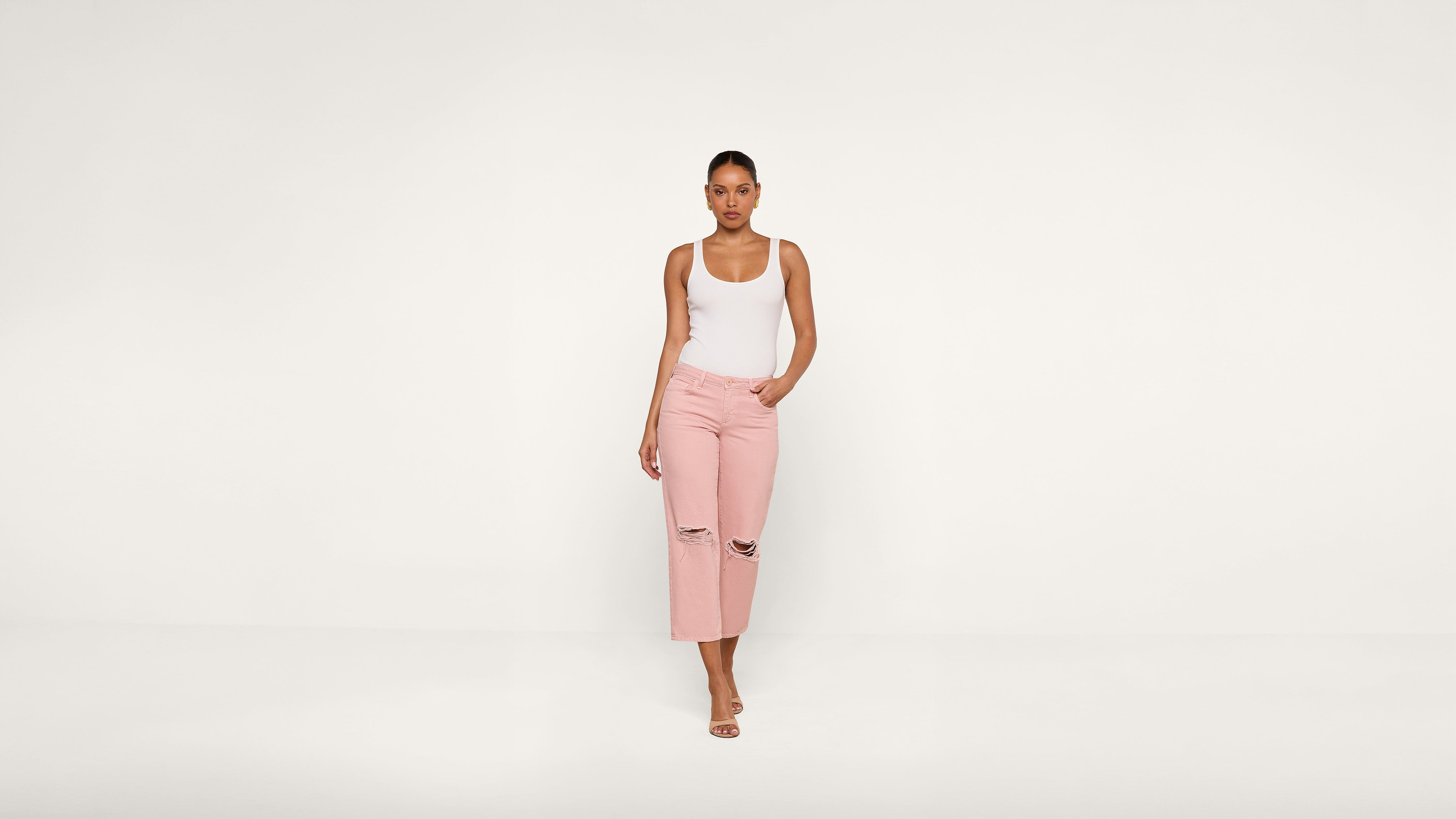A woman stands against a plain white background, wearing a white tank top, light pink ripped jeans, and sandals, with one hand in her pocket.
