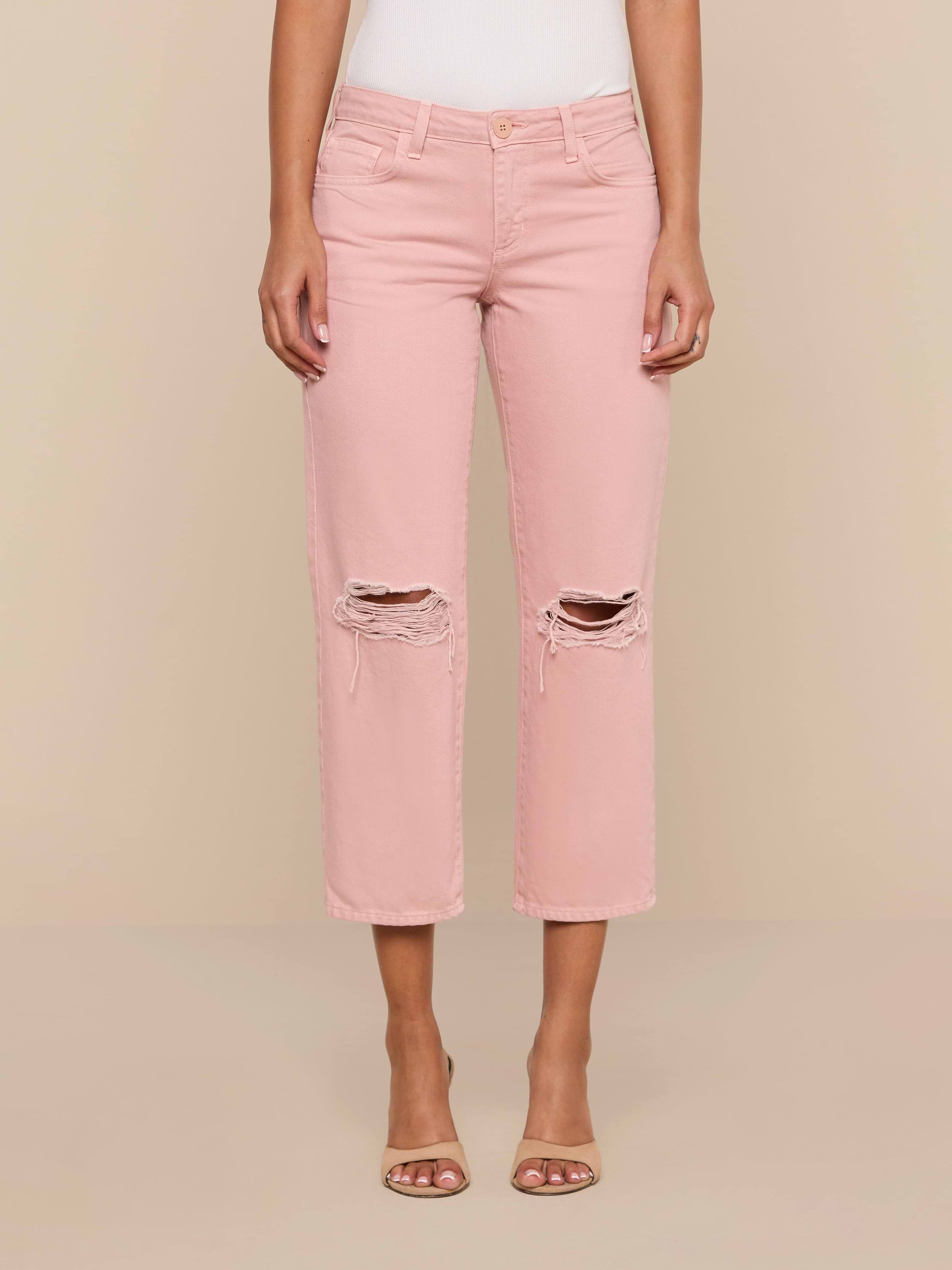 A person wearing the Blaine Cropped Stovepipe Jean in light pink with a ripped, low-rise, straight-leg fit and open-toed heels stands against a plain white background.