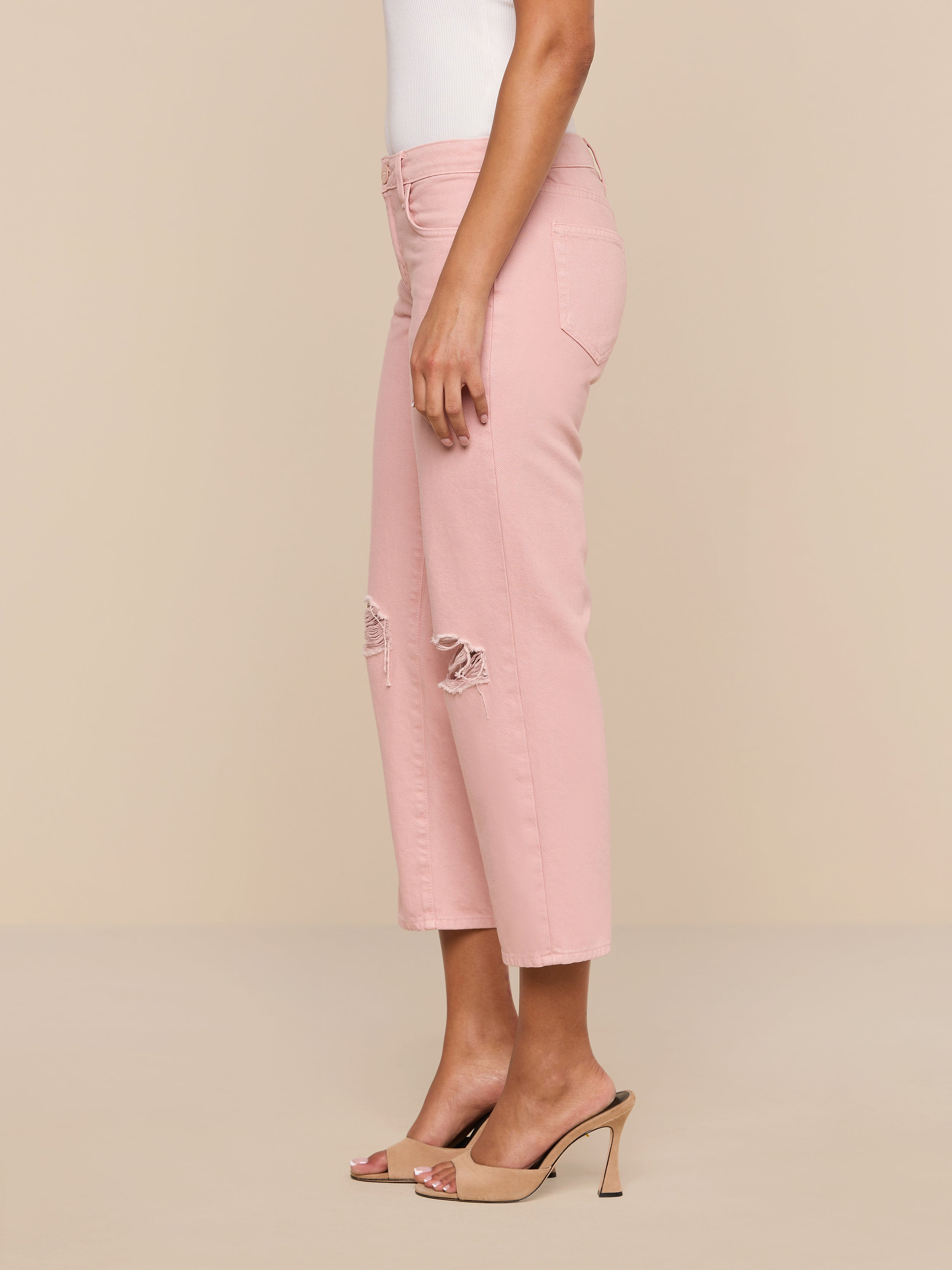 A person wearing the Blaine Cropped Stovepipe Jean in light pink with ripped knees, paired with a white sleeveless top and beige high-heeled sandals, stands sideways against a plain white background.