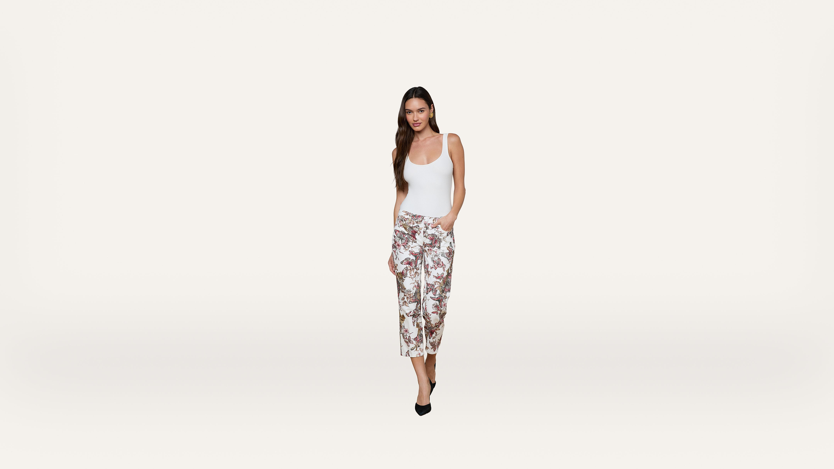 A woman in a white tank top and floral wide-leg pants stands against a plain white background, with one hand in her pocket and wearing black heels.