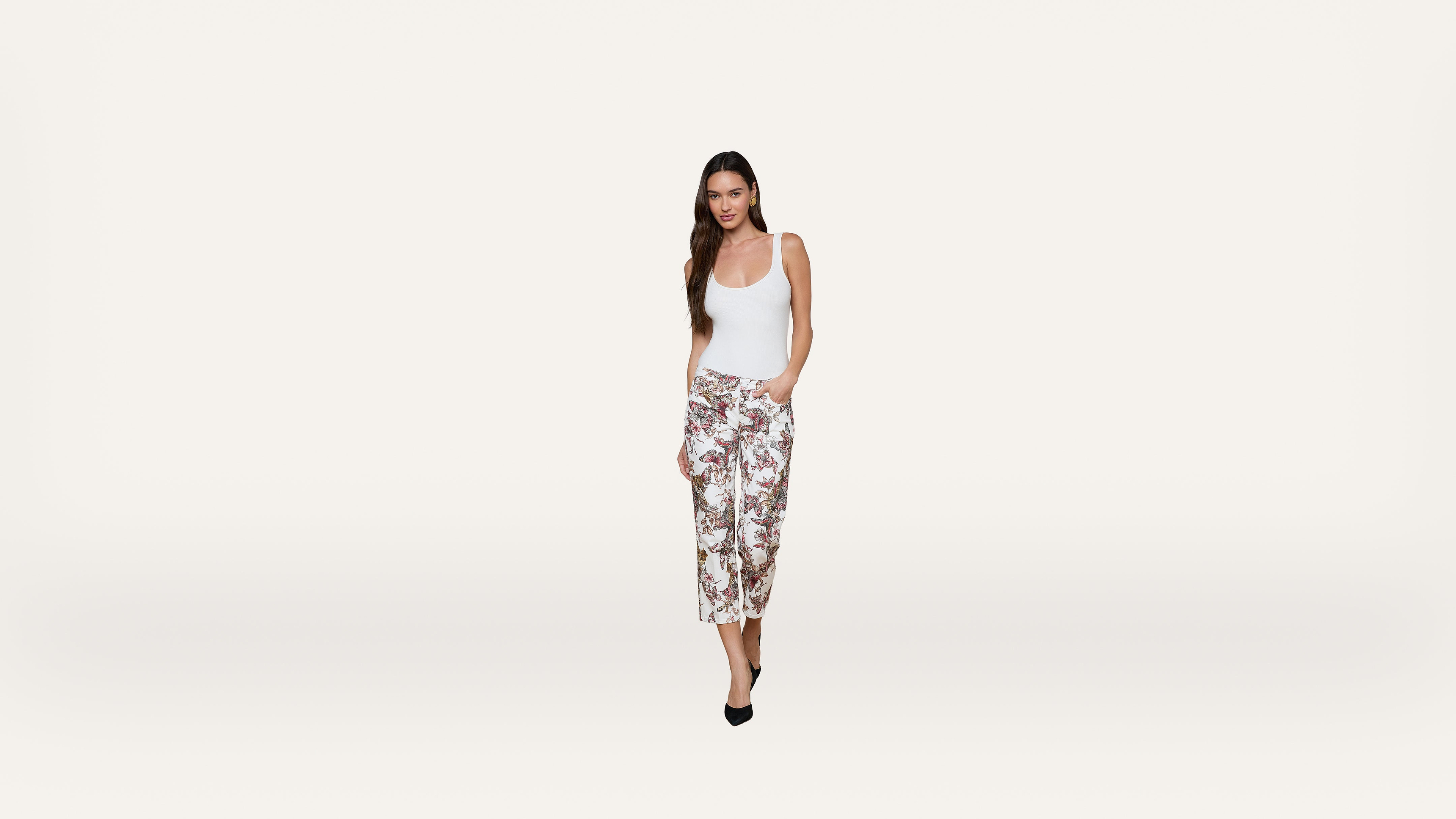 A woman in a white tank top and floral wide-leg pants stands against a plain white background, with one hand in her pocket and wearing black heels.