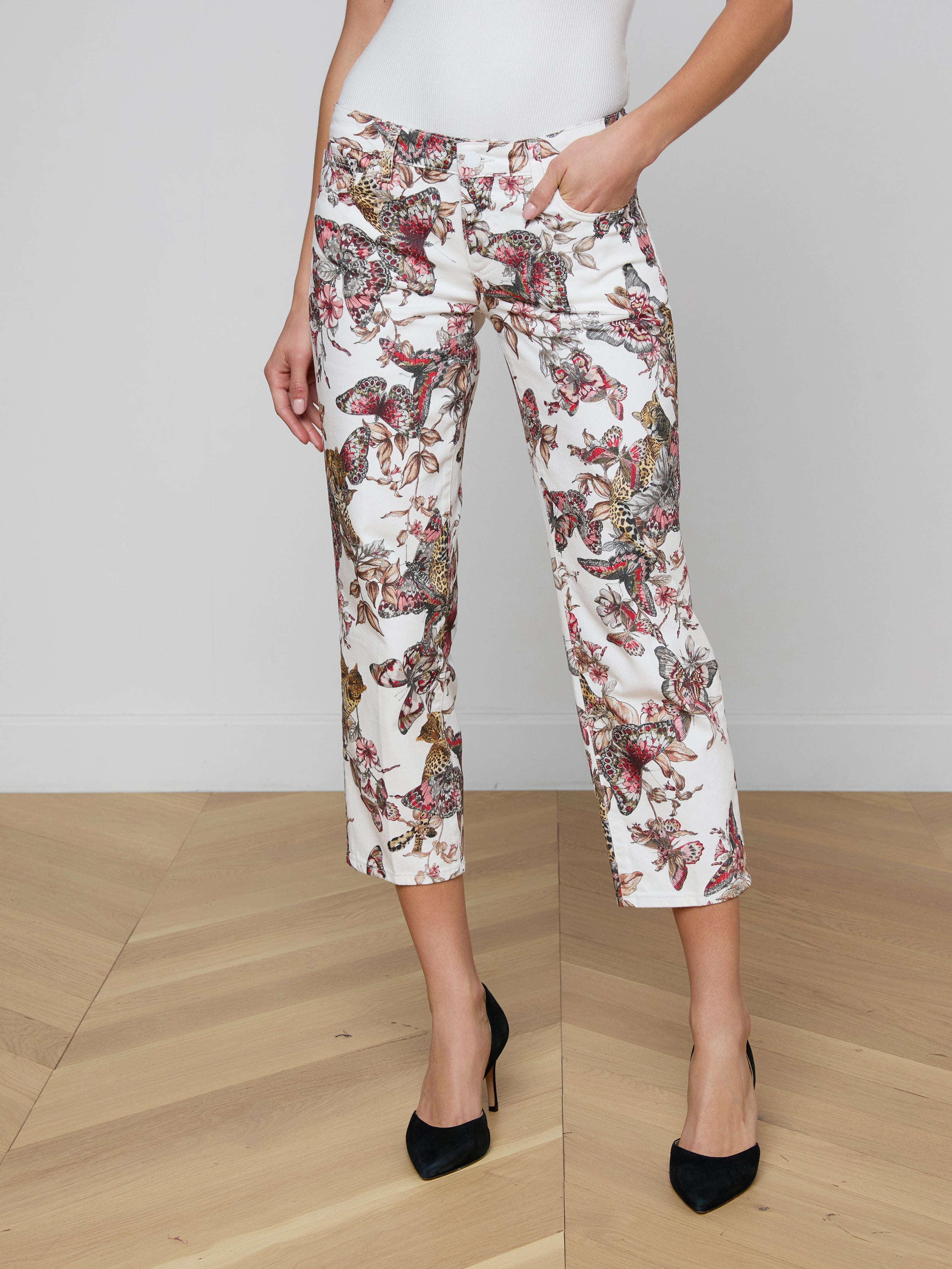 A woman models the Blaine Cropped Stovepipe Jean, featuring a colorful floral and butterfly print on white straight-leg cropped pants. She pairs them with a white top and black high-heeled shoes against a plain light background.