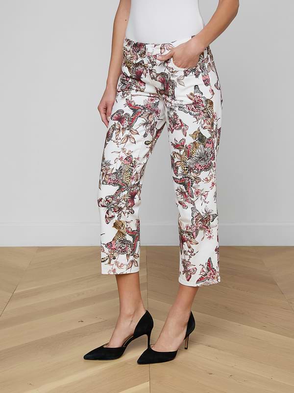A woman stands against a plain background wearing the Blaine Cropped Stovepipe Jean—white, straight-leg, ankle-length jeans featuring a colorful butterfly print—paired with a white top and black high heels, with one hand in her pocket.