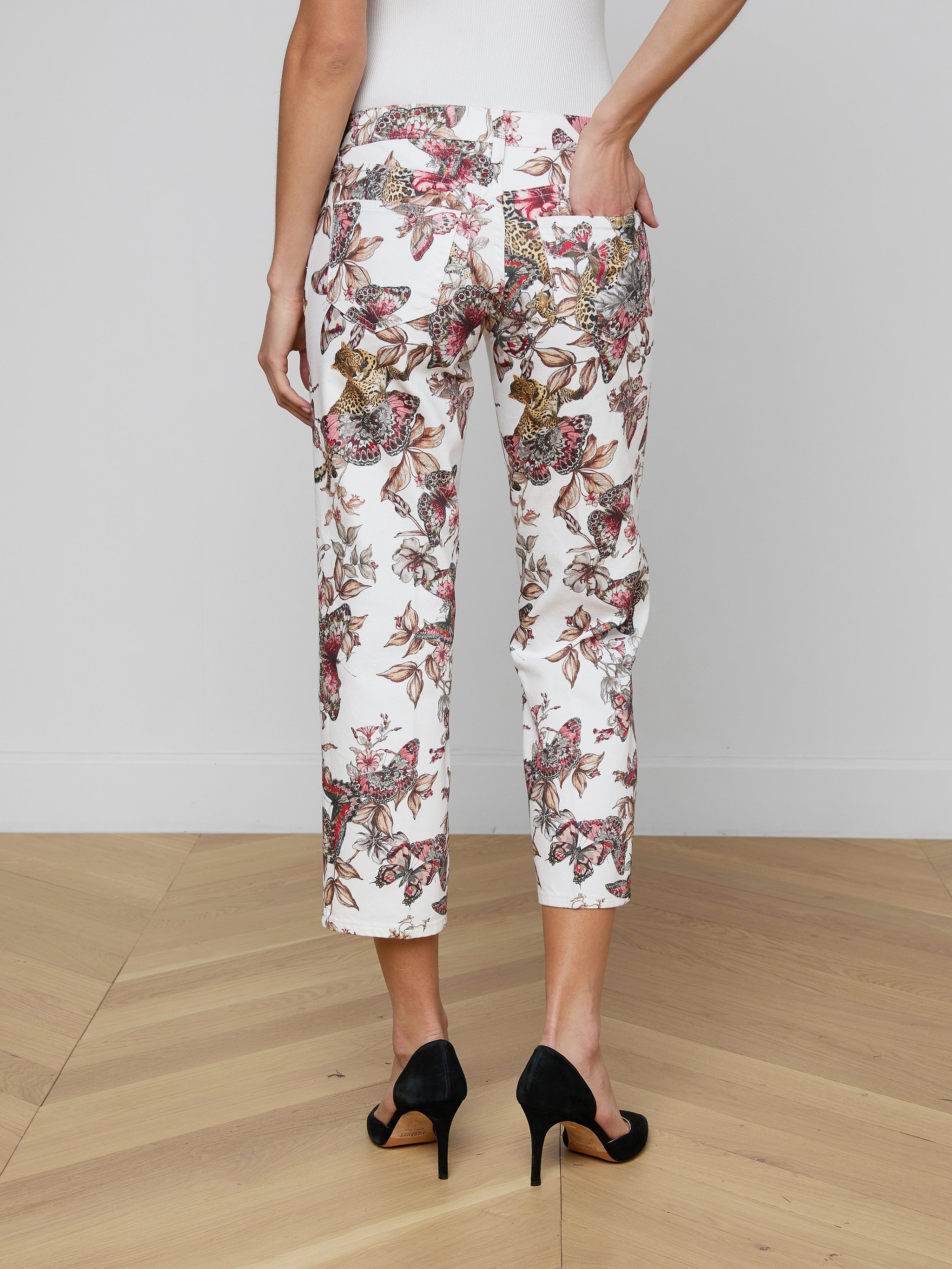 A person is seen from the back wearing the Blaine Cropped Stovepipe Jean in white with a floral print, styled with black high heels and one hand in a pocket, against a plain light background.
