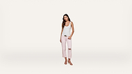 A woman stands against a plain white background, wearing a white tank top, light pink ripped jeans, and sandals. She has long brown hair and is looking at the camera with a neutral expression.