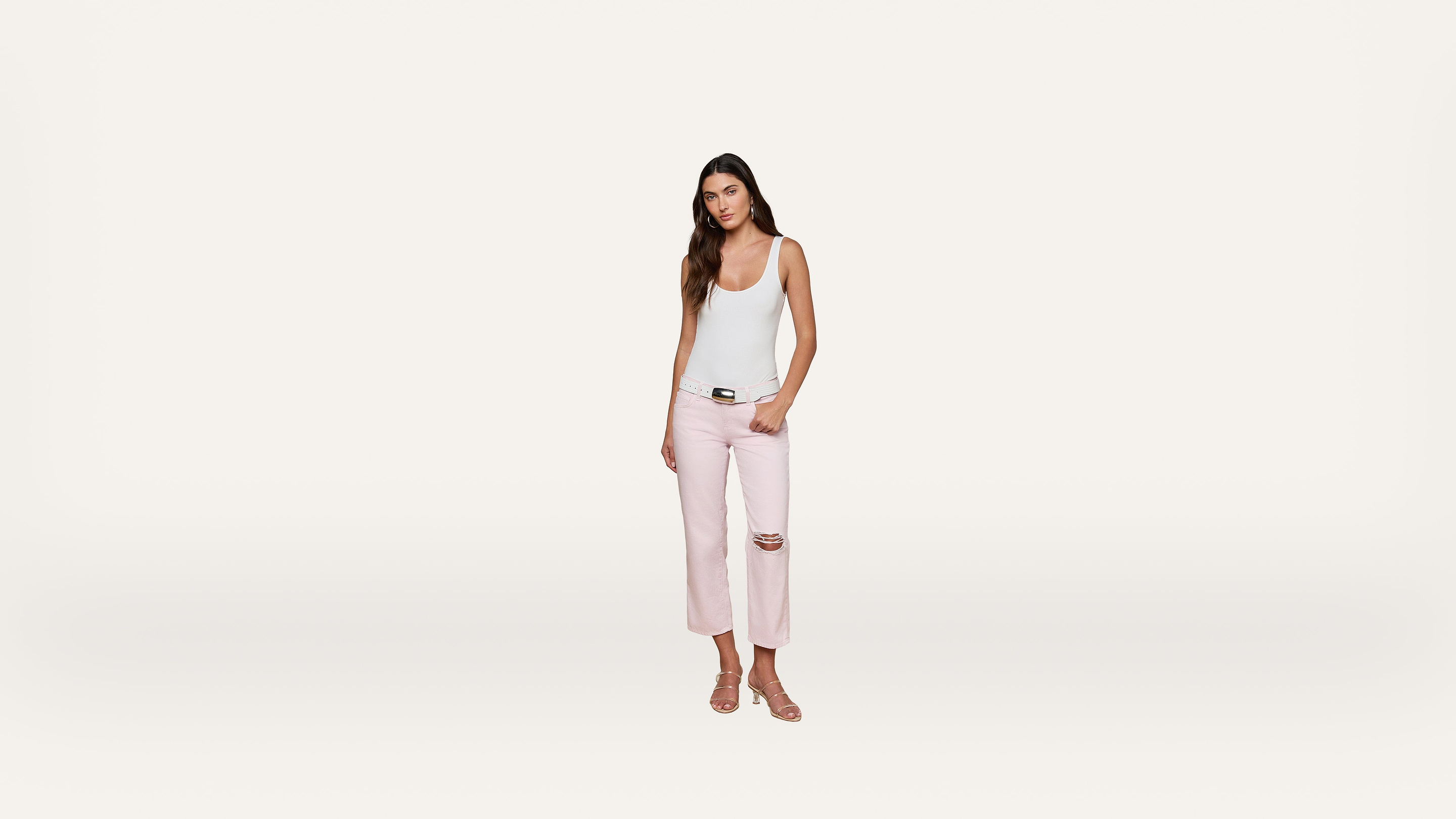 A woman stands against a plain white background, wearing a white tank top, light pink ripped jeans, and sandals. She has long brown hair and is looking at the camera with a neutral expression.