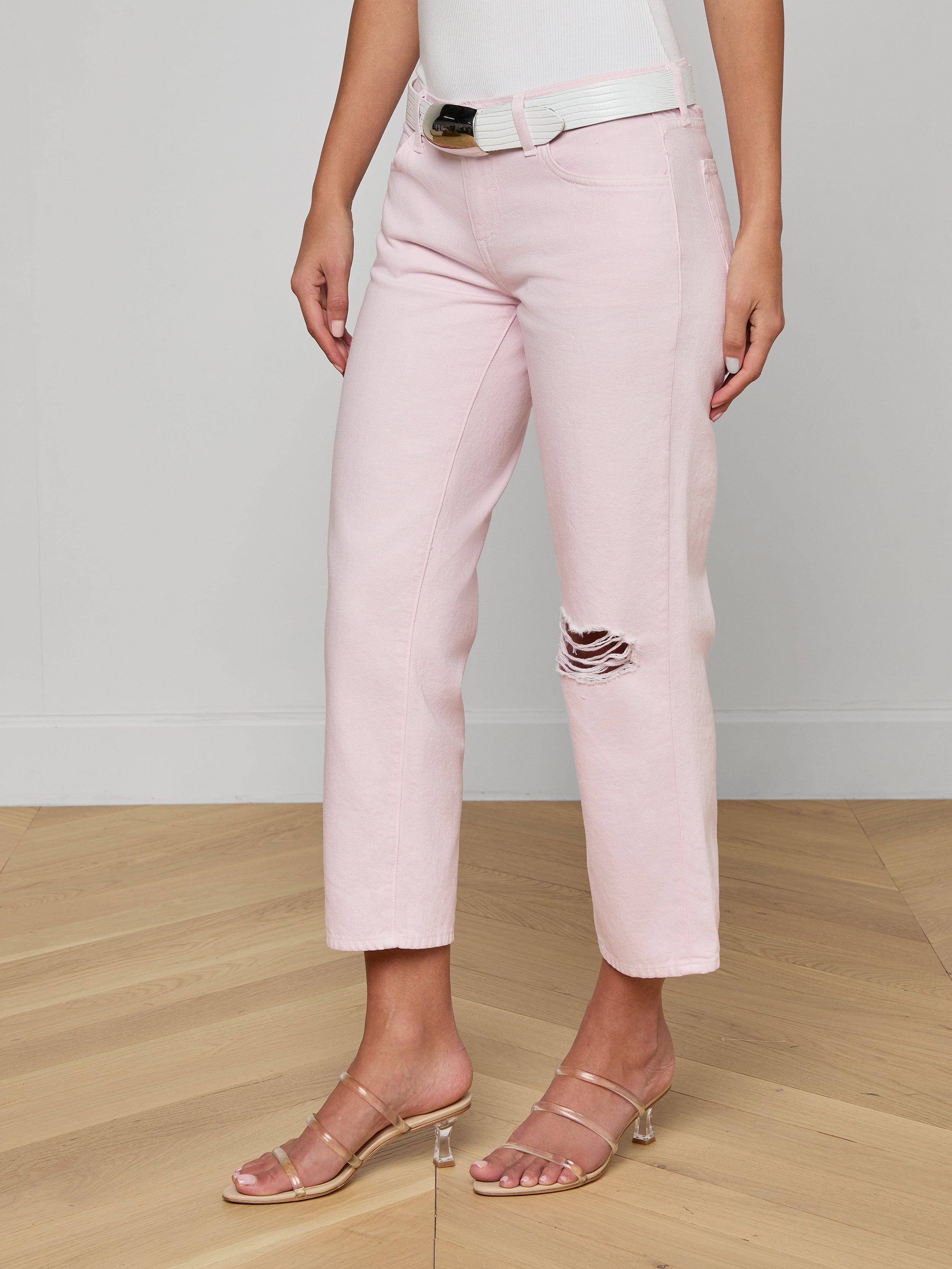 A woman wears the Blaine Cropped Stovepipe Jean in pale pink, styled with a white belt, white tank top, and strappy nude heels. The jeans have a single ripped detail on one knee. The background is plain and off-white.