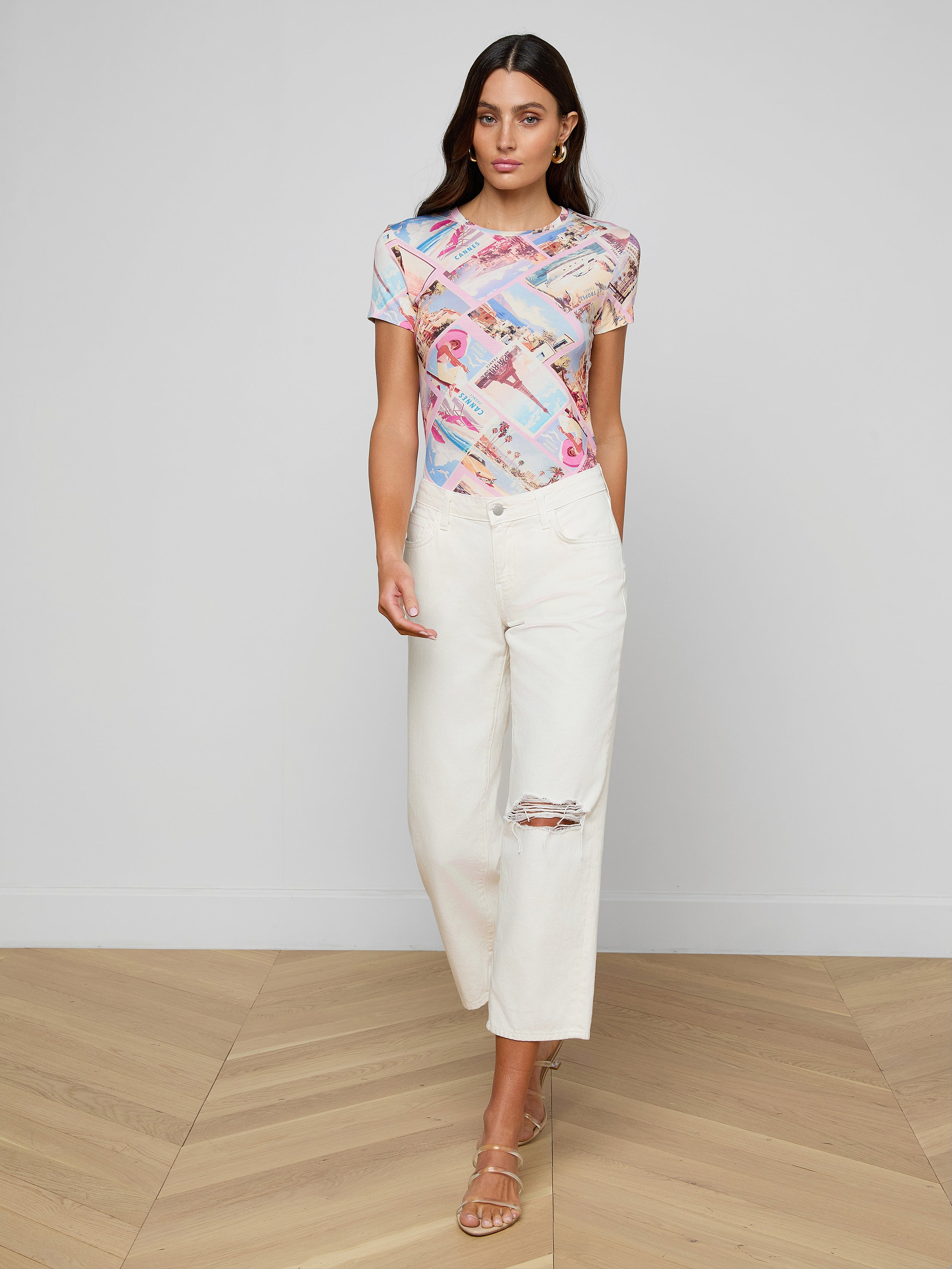 A woman wearing a colorful graphic t-shirt, Blaine Cropped Stovepipe Jeans made from all-cotton denim, and beige heeled sandals stands against a plain white background.