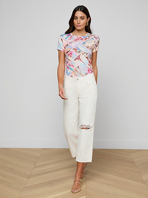 A woman stands indoors on a wooden floor, wearing a pastel-patterned short-sleeve top, beige strappy sandals, and the Blaine Cropped Stovepipe Jean—an off-white, wide-leg style with a knee rip. Her long hair is loose and she has a neutral expression.