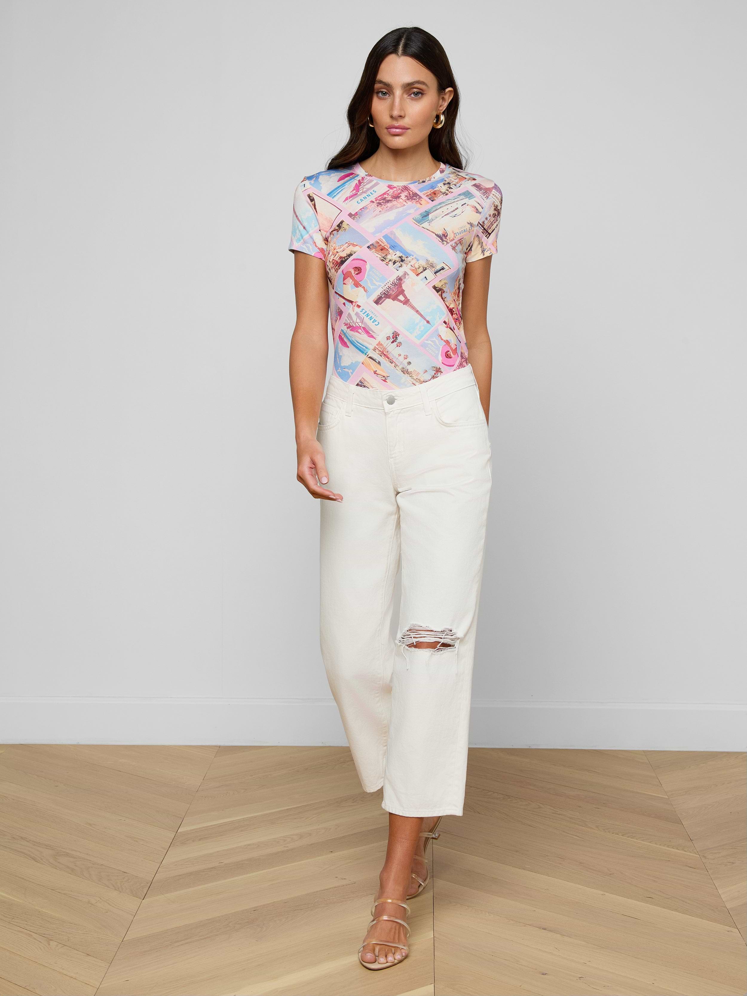A woman wearing a colorful graphic t-shirt, Blaine Cropped Stovepipe Jeans made from all-cotton denim, and beige heeled sandals stands against a plain white background.