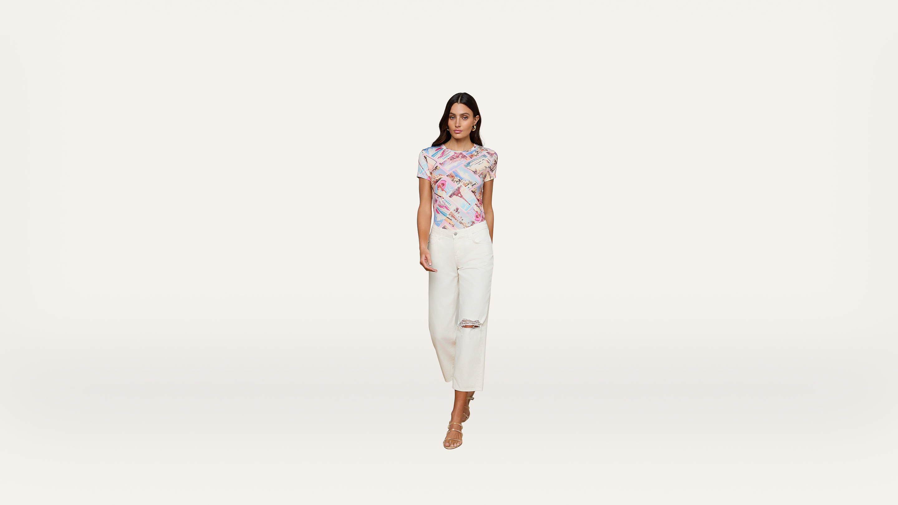 A woman stands against a white background wearing a colorful, fitted top, white ripped jeans, and nude heels. She faces forward with a neutral expression.